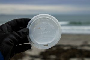 Gloved hand holding up a compostable lid with the ocean in the background.