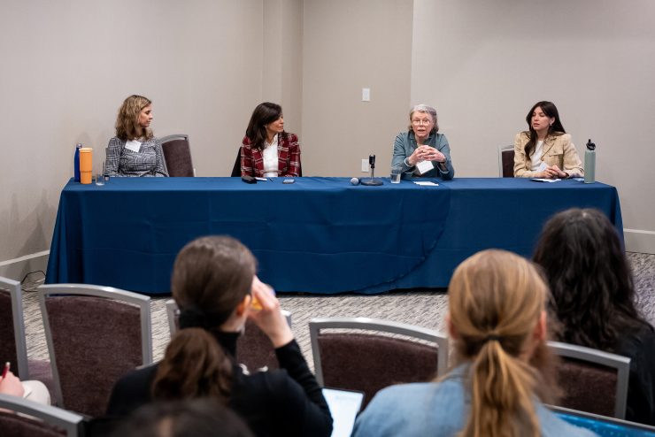 4 panelists sit behind a table as audience members watch one of them speaking.