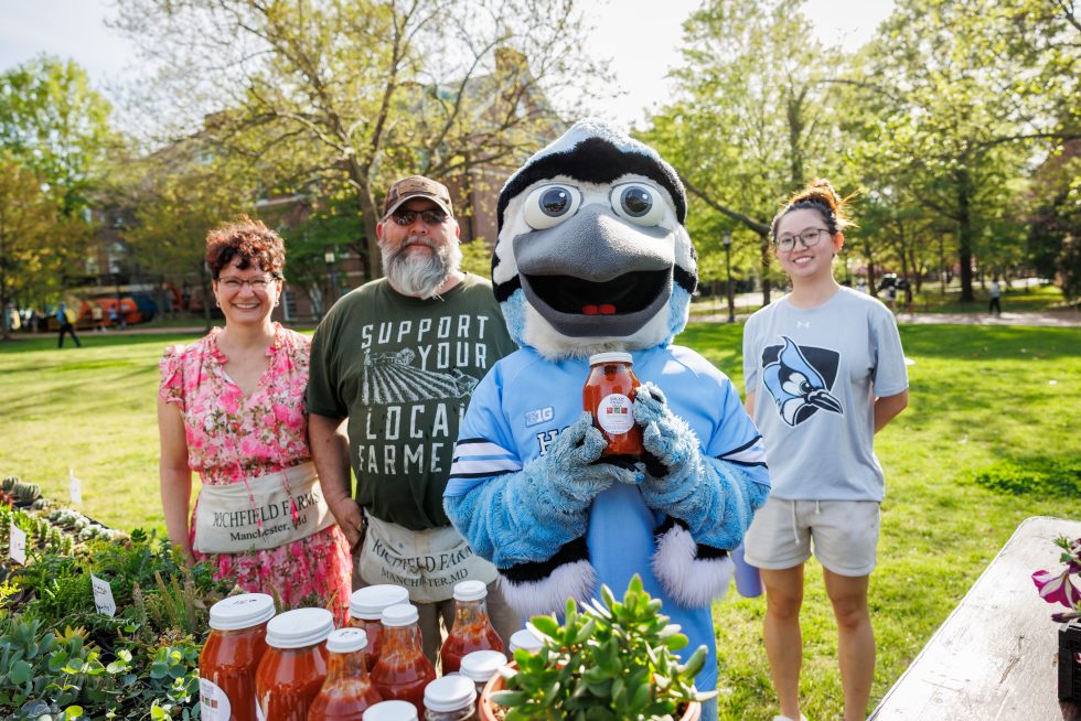 Johns Hopkins' mascot Jay holds up jar at local farmstand event on campus.