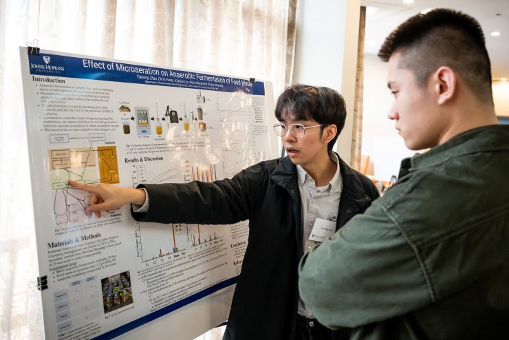 A researcher and event attendee talk while standing in front of an academic research poster.